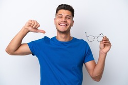 Young caucasian man holding glasses isolated on white background proud and self-satisfied