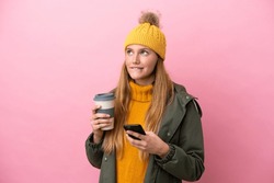 Young blonde woman wearing winter jacket isolated on pink background holding coffee to take away and a mobile while thinking something