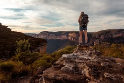Female hiker stands on a rock carrying a large backpack ... beyond her is views to mountains with sheer cliffs  and valley