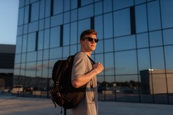 Portrait of handsome tourist hipster man in sunglasses with bag handy, standing guy near the building in downtown. Calm relaxing moment, summer vacation concept