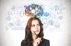 Portrait of thoughtful young businesswoman with pencil and long wavy hair standing near concrete wall with colorful brain sketch and business icons drawn on it. Concept of planning