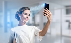 Businesswoman wearing casual t-shirt is checking her personal data. Scanning her face by smart phone to unlock it. Digital connections. Concept of data protection. Blurred office on background