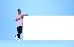 African American young man finger pointing at mockup huge poster on light blue background. Blank white canvas. Concept of presentation and ad