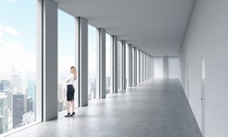 A woman in formal clothes is looking out the window. Empty modern bright clean interior of an open space office. Huge panoramic windows with New York view.