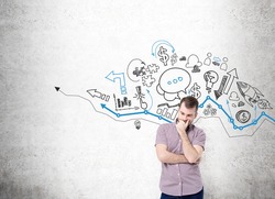 young man with hand to the chin thinking and standing in front of a concrete wall with many different business icons drawn on it over a graph. Concept of business development.