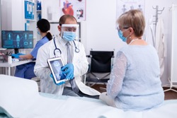 Doctor using tablet pc during examination of x-ray for older patient in hospital room and wearing face mask against coronavirus pandemic. Medical examination for infections, disease and diagnosis.
