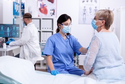 Nurse with face mask listening patient heart using stethoscope in hospital examination room wearing protection gear. Medical examination for infections, disease and diagnosis.