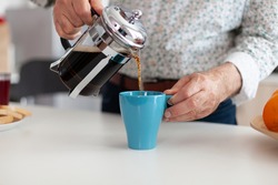 Close up of senior man pouring hot coffee from french press in kitchen during breakfast. Elderly person in the morning enjoying fresh brown cafe espresso cup caffeine from vintage mug, filter relax