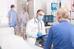 Doctor wearing visor as safety precaution in the course of consultation of senior man. Physician practitioner in the course of private modern clinic appointment during coronavirus pandemic.