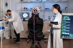 Back view of woman wearing headset for brain scan in neurology laboratory, neuro science. Nervous system analysed by team of scientists. Medical scientist looking through microscope in background