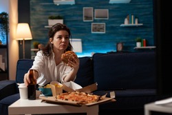 Portrait of woman holding delicious buger eating takeaway delivery food watching entertainment movie on television. Caucasian female sitting on couch tasting tasty junk-food enjoying night