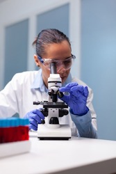 Microbiologist woman looking into medical microscope analyzing virus disease results discovering pharmacology infection. Researcher pharmacist examining bacteriology test experiment in laboratory