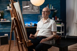 Caucasian senior man working as artist with canvas and easel for drawing masterpiece with pencil. Older artist sitting in artwork studio creating fine art as creative hobby indoors