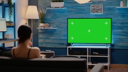 Caucasian woman looking at green screen television sitting on couch at home. Young person using chroma key for isolated background display and mockup media template while eating popcorn