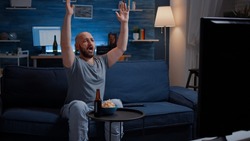 Football fan watching sport game supporting team raising hands after winning the competition, eating popcorn. Excited man celebrating good scoring of championship in living room drinking beer