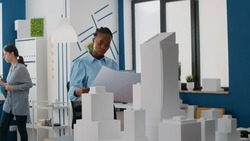 Woman builder analyzing blueprints plans to design layout of construction site and building model. Architect using maquette and property structure to build urban real estate project.