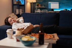 Person sleeping on sofa after drinking beer and eating home delivery pizza in front of television in living room. Tired woman falling asleep couch after large fast food takeout meal.