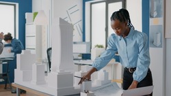 African american architect analyzing blueprints plan and building model at work. Woman engineer working on construction layout and architecture design to develop urban strategy.