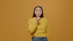 Secretive person showing silence sign on camera, covering mouth with index finger in studio. Mysterious woman doing silent and mute gesture, no noise pose over yellow background. Secrecy