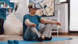 Old woman wearing vr glasses and doing meditation while sitting in lotus position on yoga mat. Senior person using virtual reality goggles to meditate for wellness and recreation.