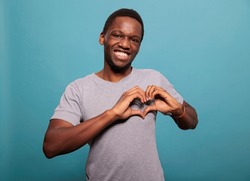Young adult doing heart shaped sign with hands in studio, expressing amorous feelings on camera. Smiling man doing romantic gesture to express love, affection and passion for valentines day.