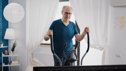 Retired man doing cardio exercise with stationary bicycle to have workout training activity. Pensioner using electric machine to cycle and do gymnastics. Old person with fitness bike.