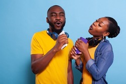 Trendy people doing karaoke with mobile phone as microphone in studio. Man and woman singing and having fun together with playlist music on headphones, enjoying mp3 sound on headset.