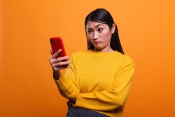 Doubtful uncertain beautiful woman wearing yellow sweater while using smartphone device on orange background. Confused clueless adult person using mobile cellphone to understand un clear instructions.