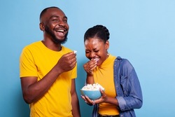 Joyful man and woman watching funny movie on television, eating popcorn in studio and laughing. Happy couple with snack looking at comedy film on tv, having fun and enjoying laugh together.