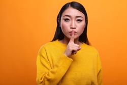 Calm attractive women wearing yellow sweater making hush gesture with forefinger on orange background. Mysterious person touching lips with index finger indicating silence and secrecy.