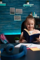 Schoolkid reader smiling while reading edcuational book studying for school literature exam sitting at desk in living room. Little child working at academic homework during home schooling