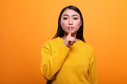 Beautiful mysterious woman wearing vibrant yellow sweater making silence gesture on orange background. Secretive person touching lips with forefinger indicating silence and secrecy.