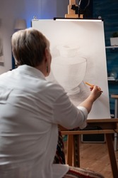 Portrait of elderly artist sketching still life using sharp pencil on paper sketching vase model in home art studio. Over shoulder view of older woman drawing orginal masterpiece in workshop.