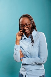 Cheerful adult person talking with friend on touchscreen cellphone device. Happy joyful young woman smiling heartily while having a phone call conversation on blue background. Studio shot