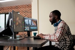 Video production house employee sitting at multi monitor workstation while editing movie frames using advanced software. African american graphic footage editor enhancing project quality.