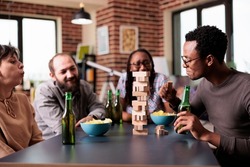 Multiethnic group of friends sitting at table in living room while playing together society games. Diverse people sitting at home while enjoying party game with wooden blocks and tower structure.