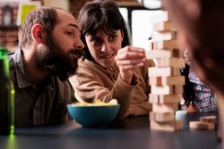 Focused people sitting at table in living room while playing with wood blocks. Skilled concentrated woman carefully removing wooden cube from tower structure while enjoying fun leisure time activity.