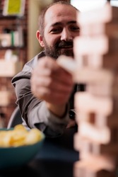 Selective focus of smiling man pulling wooden cube from blocks tower structure. Happy person enjoying friends company while playing society games together at home in living room.