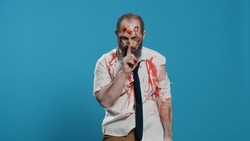 Silent creepy zombie making shush secrecy hand gesture on blue background. Dangerous walking dead corpse with deep and bloody wounds being confidential while smirking at camera.