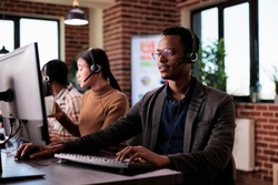 African american helpline employee working at call center reception with multiple monitors. Male operator using telecommunication to help clients at customer service support, remote network.