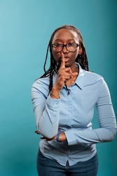 Attractive serious woman making silence gesture by putting forefinger over lips on blue background. Young adult person shushing people indicating secrecy and confidentiality.