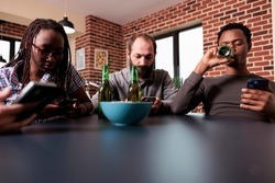 Diverse group of friends sitting in living room with smartphones. Multiracial people at home surfing webpages on modern touchscreen electronic devices while having beer and snacks.
