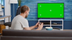 Man holding wireless controller playing console video game on green screen tv while sitting on sofa in modern living room. Gamer relaxing on couch enjoying online gaming on croma key display.