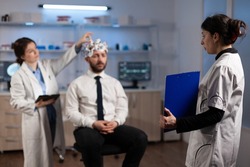Researcher woman holding clipboard with neuroscience information on it while neurologist doctor adjusting eeg headset monitoring brain activity of man patient. Specialist analyzing nervous system