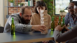 Positive people having fun with society game in living room, playing with wooden tower blocks. Men and women enjoying game with building square pieces on structure for entertainment.