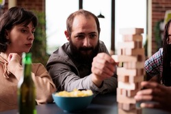 Focused man pulling wood block from tower structure while playing society games with friends. Multiethnic group of people enjoying fun leisure activity together while sitting at home in living room.