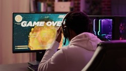 Over shoulder view of gamer feeling unhappy after losing difficult level in online space shooter on gaming pc at home. Man playing internet multiplayer game disappointed after failing competition.