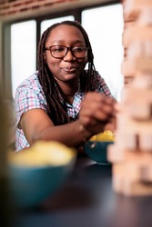 Joyful woman carefully removing wood block from wooden tower structure while sitting at table. Focused smiling person enjoying playing society games with friends at home in living room.