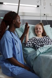 Childcare healthcare facility staff high fiving ill kid under medical treatment. Nurse doing high five gesture with sick girl resting in hospital pediatric ward patient bed.