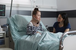 Neck injured hospitalized girl with cervical collar resting in hospital pediatric ward. Unwell little kid suffering from car accident sitting in patient bed while mother comforts her.